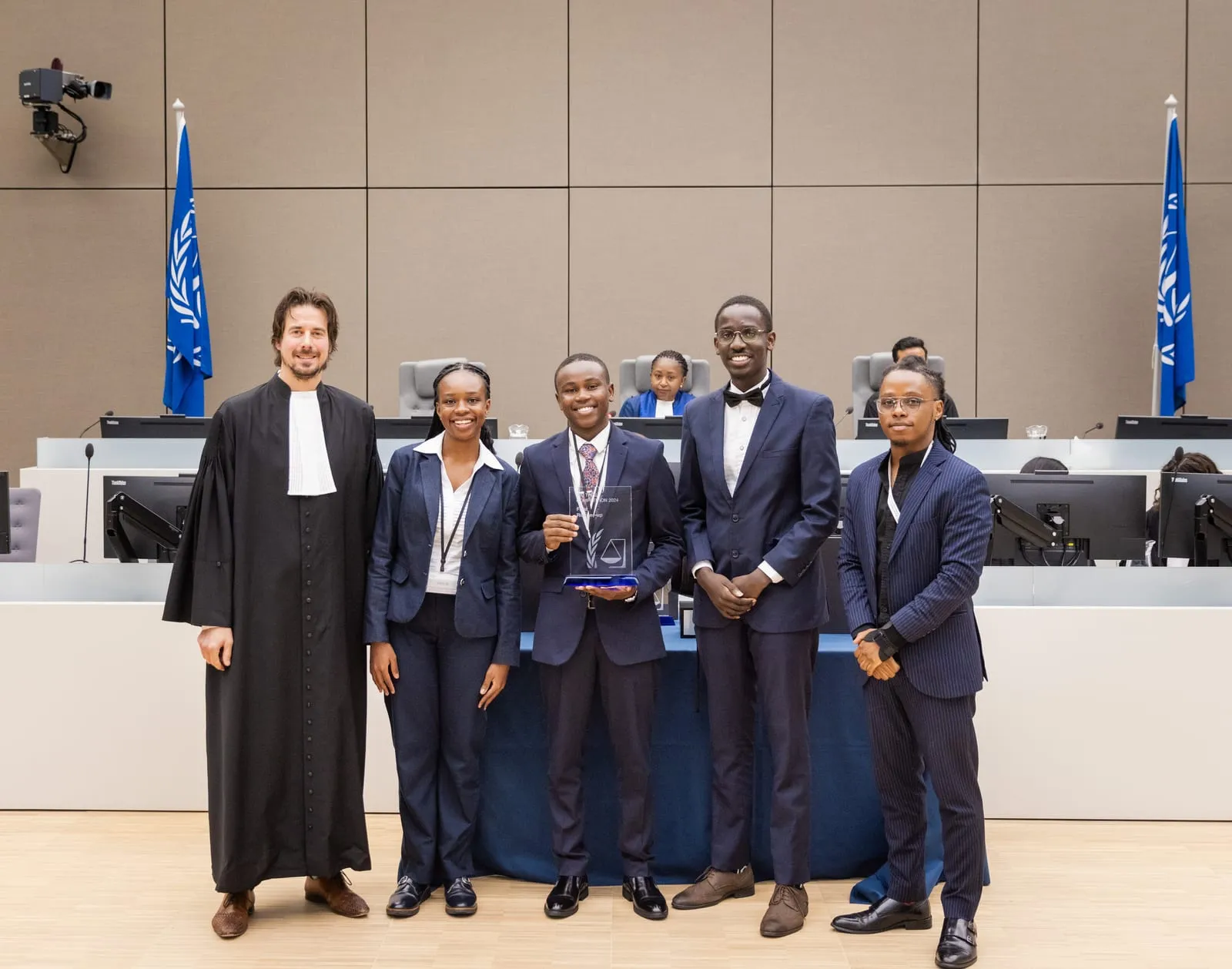 A judge with a team receiving their award, the ICC emblem flags behind, at the 2024 ICC Moot Court Competition Final