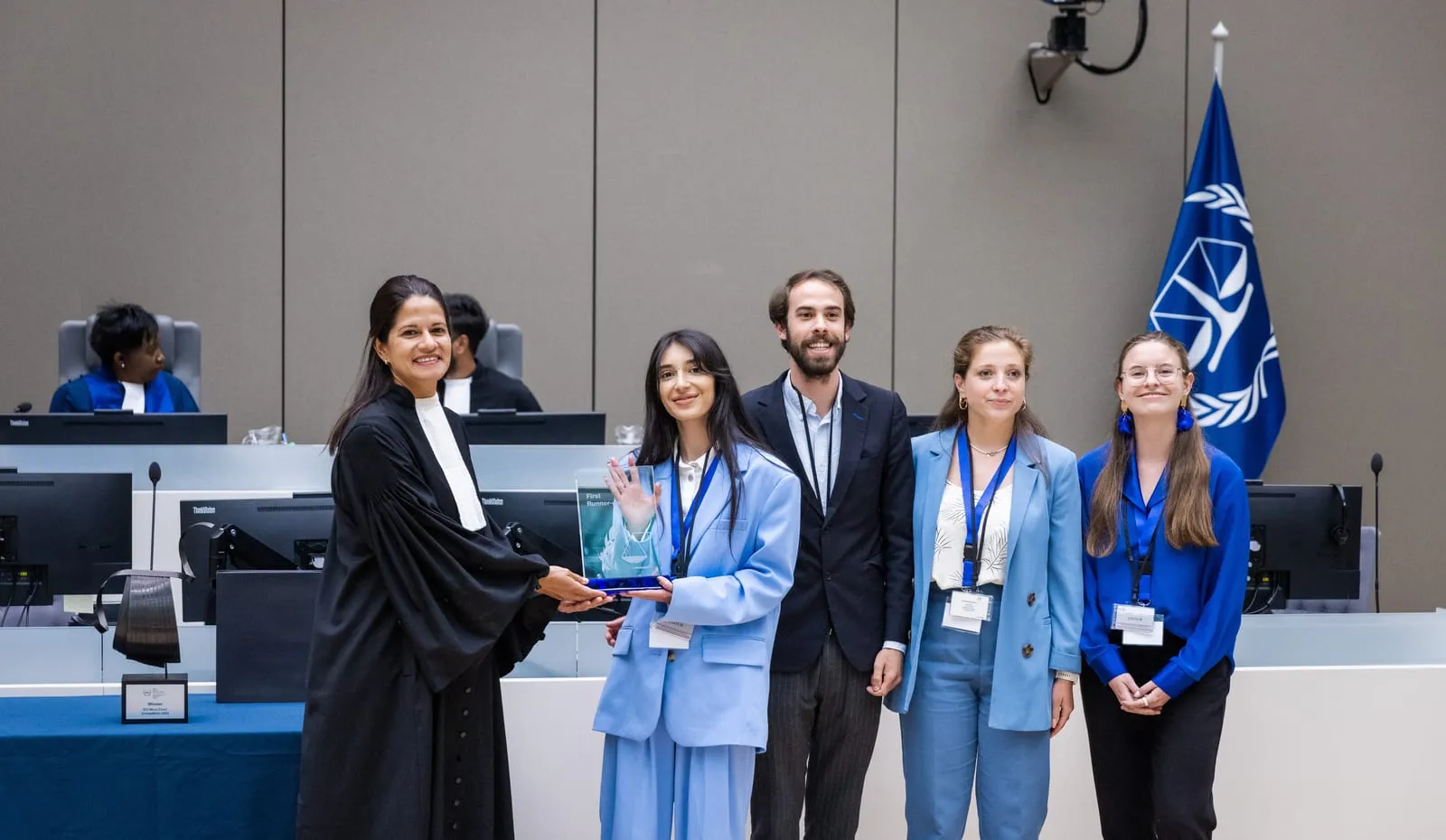 A judge presenting the first runner-up award to a team, the ICC emblem flag behind them, at the 2023 ICC Moot Court Competition Final